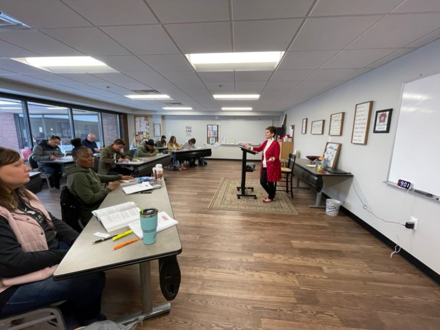 A room with people sitting at tables and standing in front of windows.